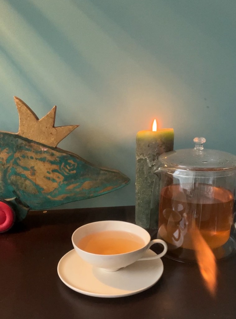 A cozy scene featuring a cup of tea on a saucer, a glass teapot filled with brewed tea, a green candle with a flame, and a decorative fish figure with a crown set against a teal wall.