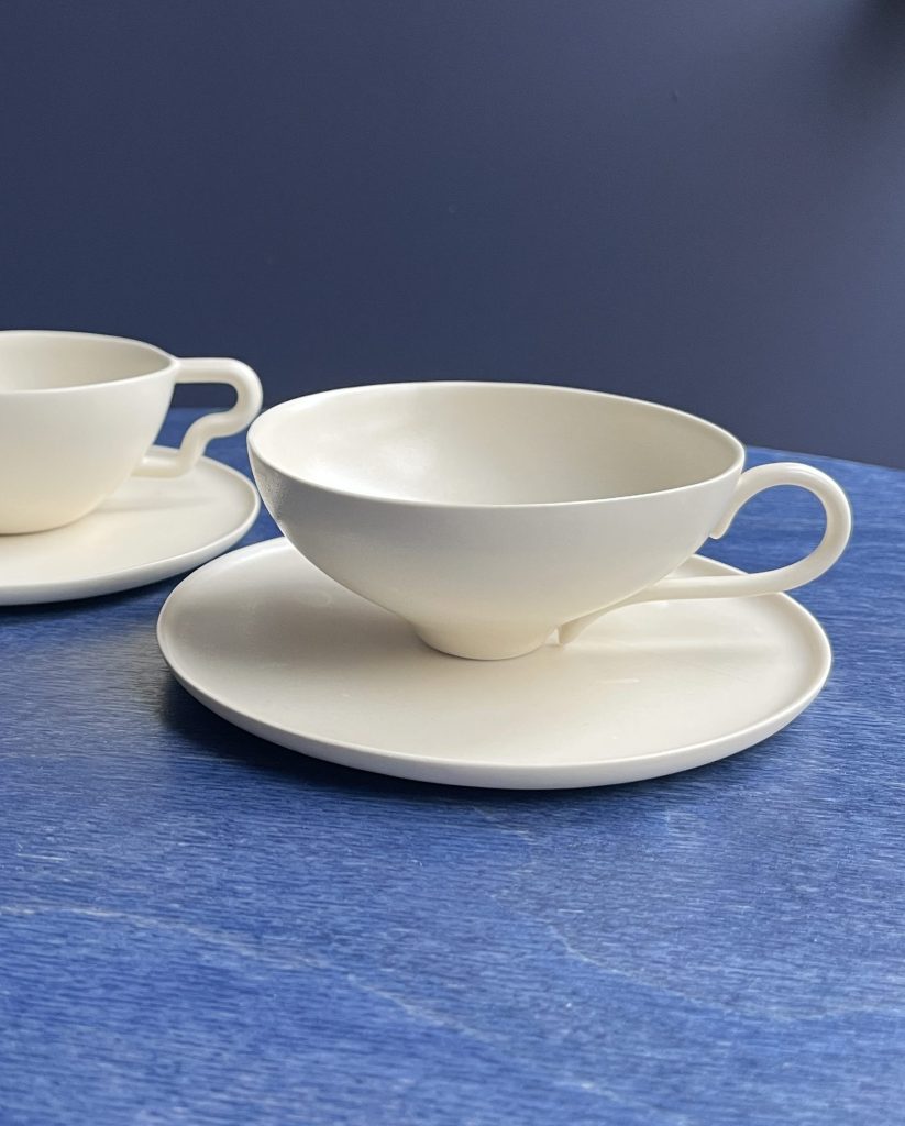 A white ceramic teacup with a matching saucer, set against a blue background. Another similar teacup is visible behind it.